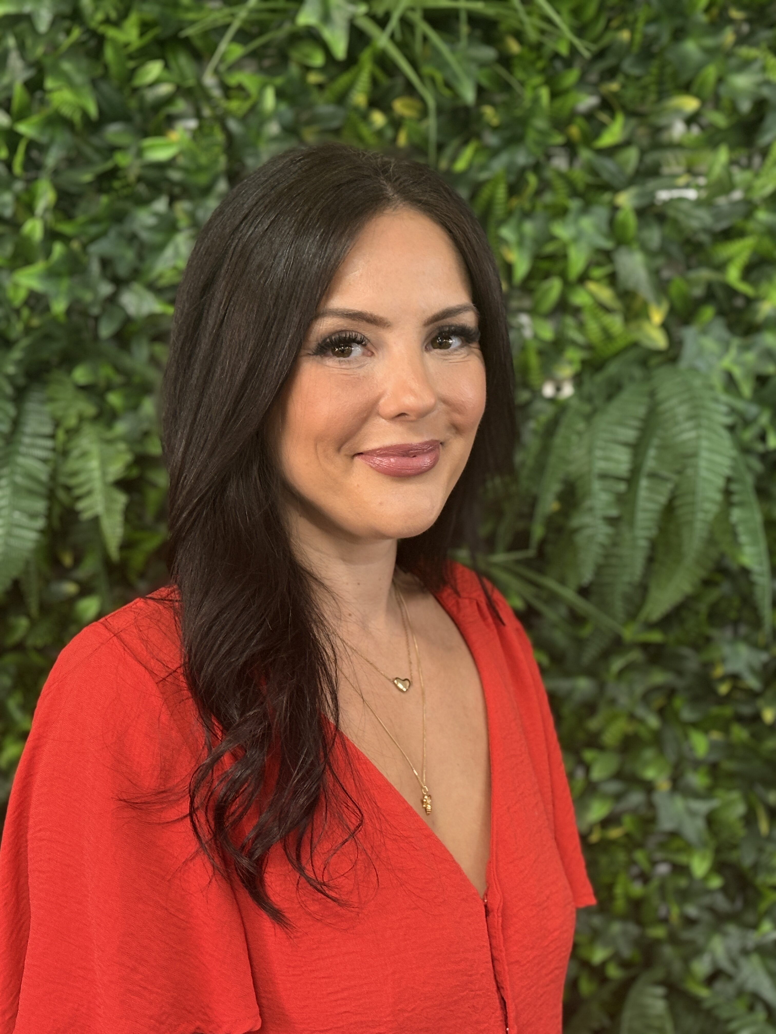 A female wearing a red top, smiling at the camera standing in front of a green foliage background.